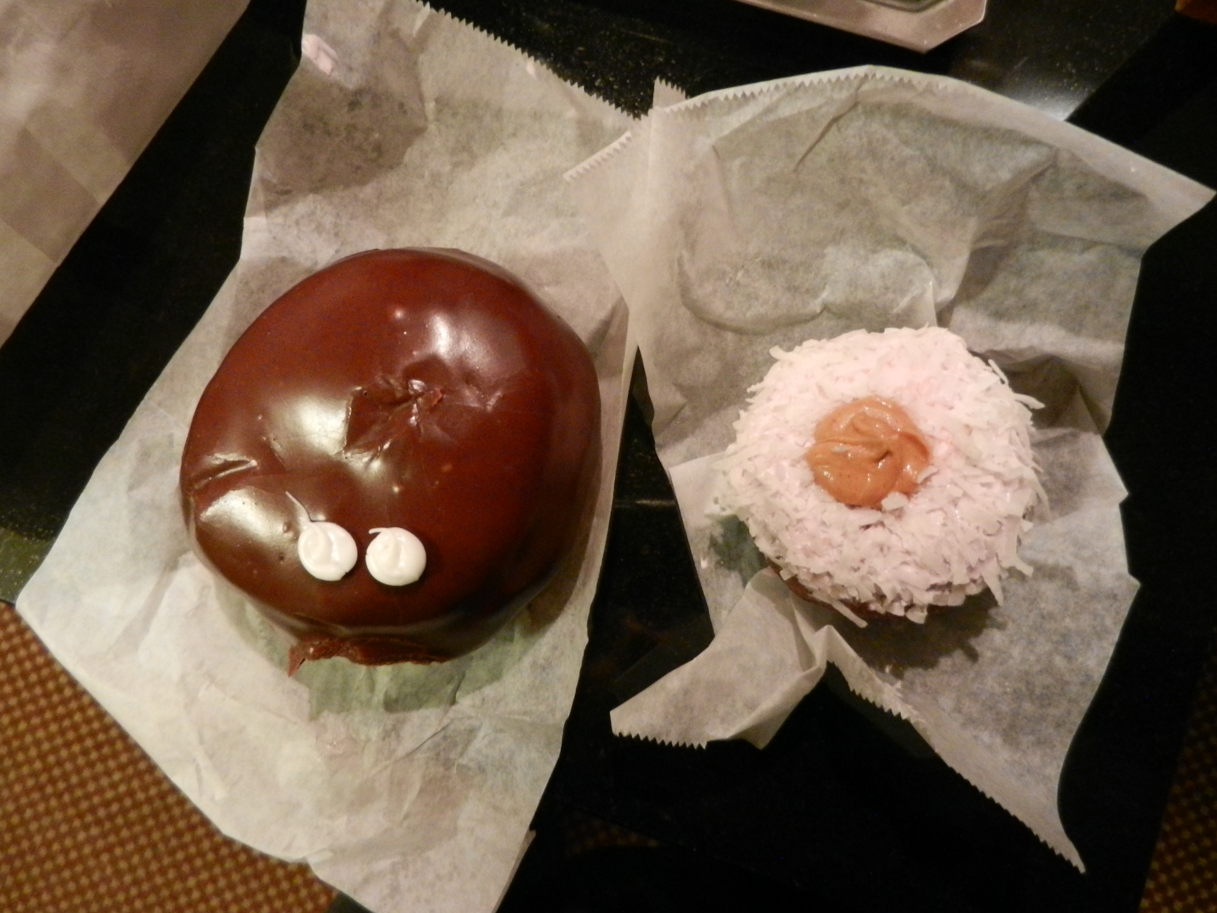Two doughnuts - on the left a donut with chocolate frosting and two white "eyes" and on the right a doughnut with pink frosting, coconut on top, and peanut butter in the middle.