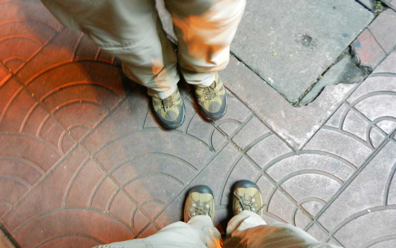 Woman's feet at the bottom, man's feet at the top of the photo - both wearing brown hiking boots and standing on a brownish-orange sidewalk depicting a half-circle pattern.