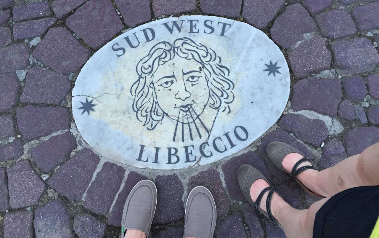 Man's feet on the left wearing tan shoes, woman's feet on the right wearing grey shoes standing on a brick surface next to a medallion depicting a head blowing air out its mouth surrounded by the words "Sud West, Libeccio".