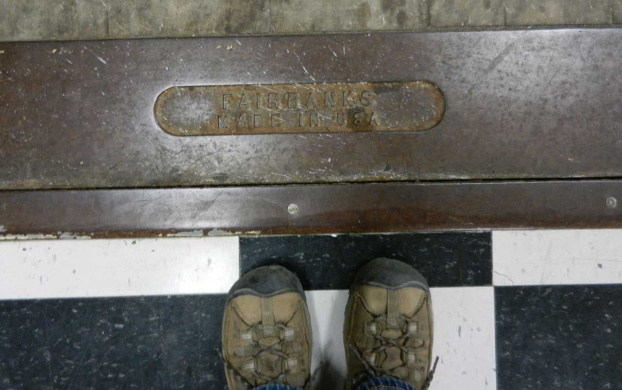 Women's feet wearing hiking boots standing on a black and white tiled floor next to the metal base of a luggage scale with a metal placard that reads "Fairbanks, Made in USA".