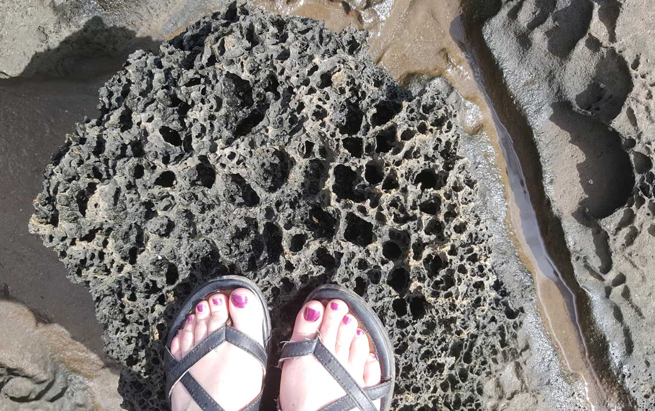Women's feet with purple toe nail polish wearing black sandals and standing on a porous black lava rock.