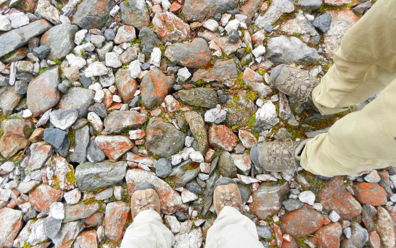 Woman's feet at the bottom, man's feet on the right - both wearing brown hiking boots - standing on grey, white, brown, and red rocks.